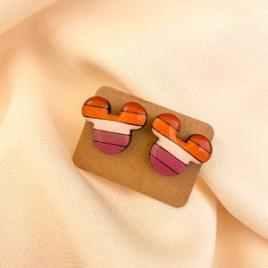 May include: A pair of colorful, striped Mickey Mouse-shaped earrings. The earrings are made of polymer clay and have a glossy finish. The colors are orange, pink, white, and black.