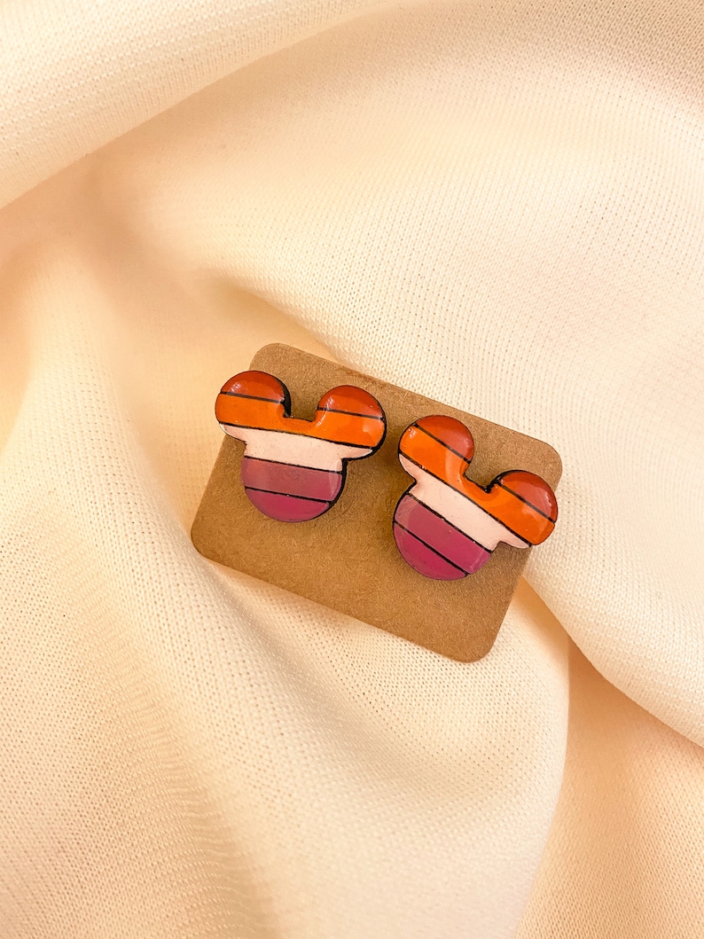 May include: A pair of colorful, striped Mickey Mouse-shaped earrings. The earrings are made of polymer clay and have a glossy finish. The colors are orange, pink, white, and black.