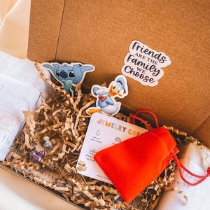 May include: A brown cardboard box filled with brown packing paper. The box contains a red pouch, two small colorful earrings, a card with the text "Jewelry Care" and three stickers. One sticker features Stitch from Lilo & Stitch, another features Donald Duck, and the third sticker says "Friends are the family we choose."