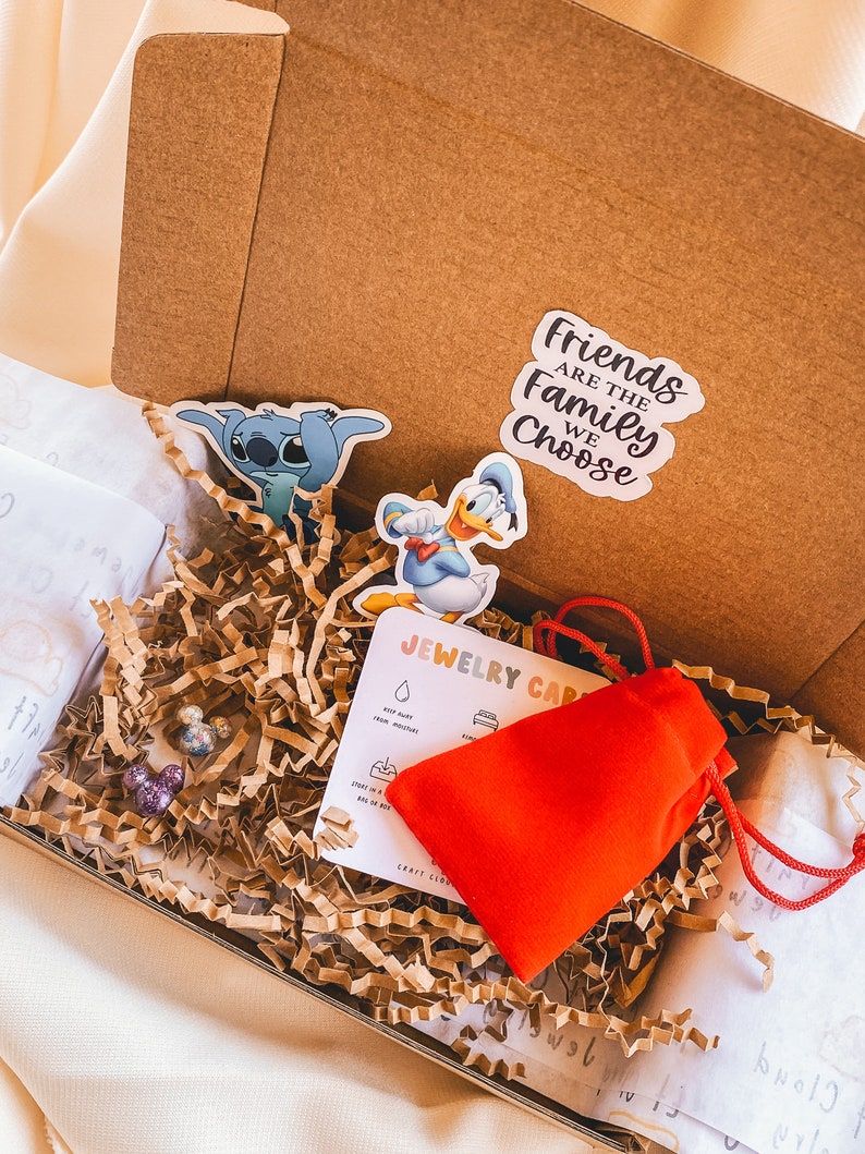 May include: A brown cardboard box filled with brown packing paper. The box contains a red pouch, two small colorful earrings, a card with the text "Jewelry Care" and three stickers. One sticker features Stitch from Lilo & Stitch, another features Donald Duck, and the third sticker says "Friends are the family we choose."