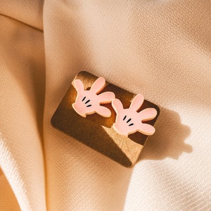 Puede incluir: Un par de pendientes de mano de dibujos animados de color rosa con detalles negros. Los pendientes est&aacute;n sobre una superficie marr&oacute;n.