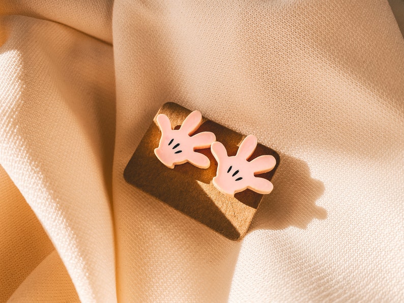 Puede incluir: Un par de pendientes de mano de dibujos animados de color rosa con detalles negros. Los pendientes est&aacute;n sobre una superficie marr&oacute;n.