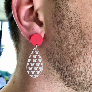 May include: A pair of dangle earrings with a red circle and a teardrop-shaped pendant. The pendant is clear and has a white Mickey Mouse pattern.