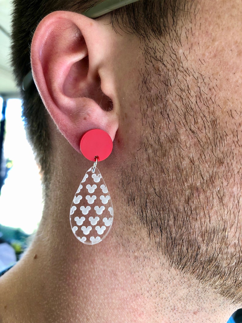 May include: A pair of dangle earrings with a red circle and a teardrop-shaped pendant. The pendant is clear and has a white Mickey Mouse pattern.