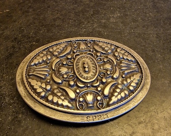 Könnte beinhalten: Ovale, silberfarbene Metallschnalle mit einem verzierten, geprägten Design. Die Schnalle zeigt florale und scrollartige Muster um ein zentrales ovales Element. Das Wort "ESPRIT" ist unten geprägt. Wahrscheinlich ein Vintage-Artikel.