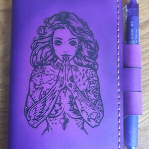 May include: A purple leather journal with a black and white engraved image of a woman with tattoos. The journal has a pen holder and a pink stitch around the edges.