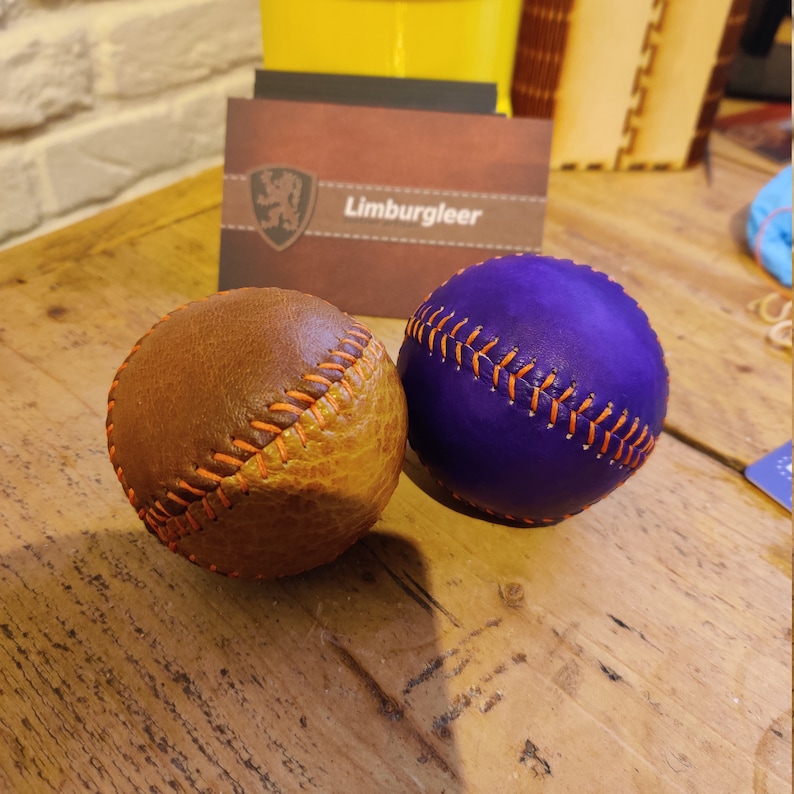 May include: Two leather baseballs, one brown and one purple, with orange stitching. The balls are on a wooden surface.