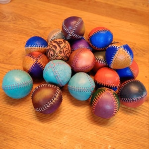 May include: A collection of 15 colorful leather balls with stitched seams. The balls are arranged in a pyramid shape on a wooden surface. The balls are various shades of blue, purple, red, brown, and tan.