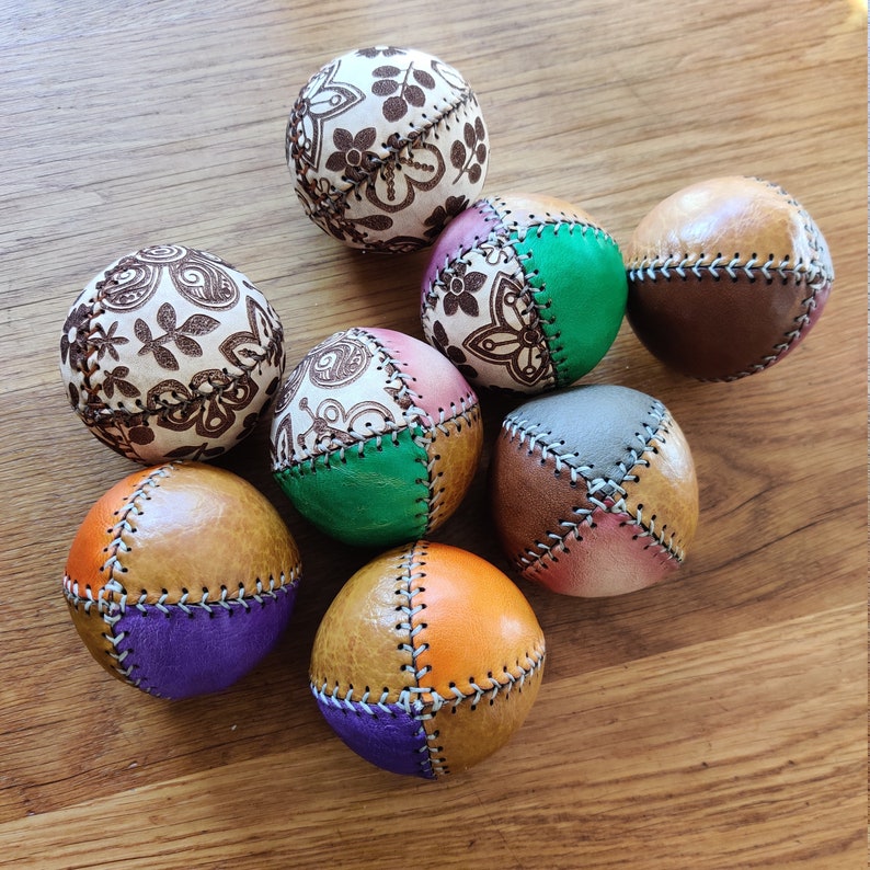 May include: Eight multi-colored leather juggling balls with different patterns and stitching. The balls are arranged in a circle on a wooden surface.