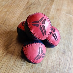 May include: Four leather juggling balls with a black and red design. Each ball has a stitched seam and a black logo of a stylized triangle with a line through it.