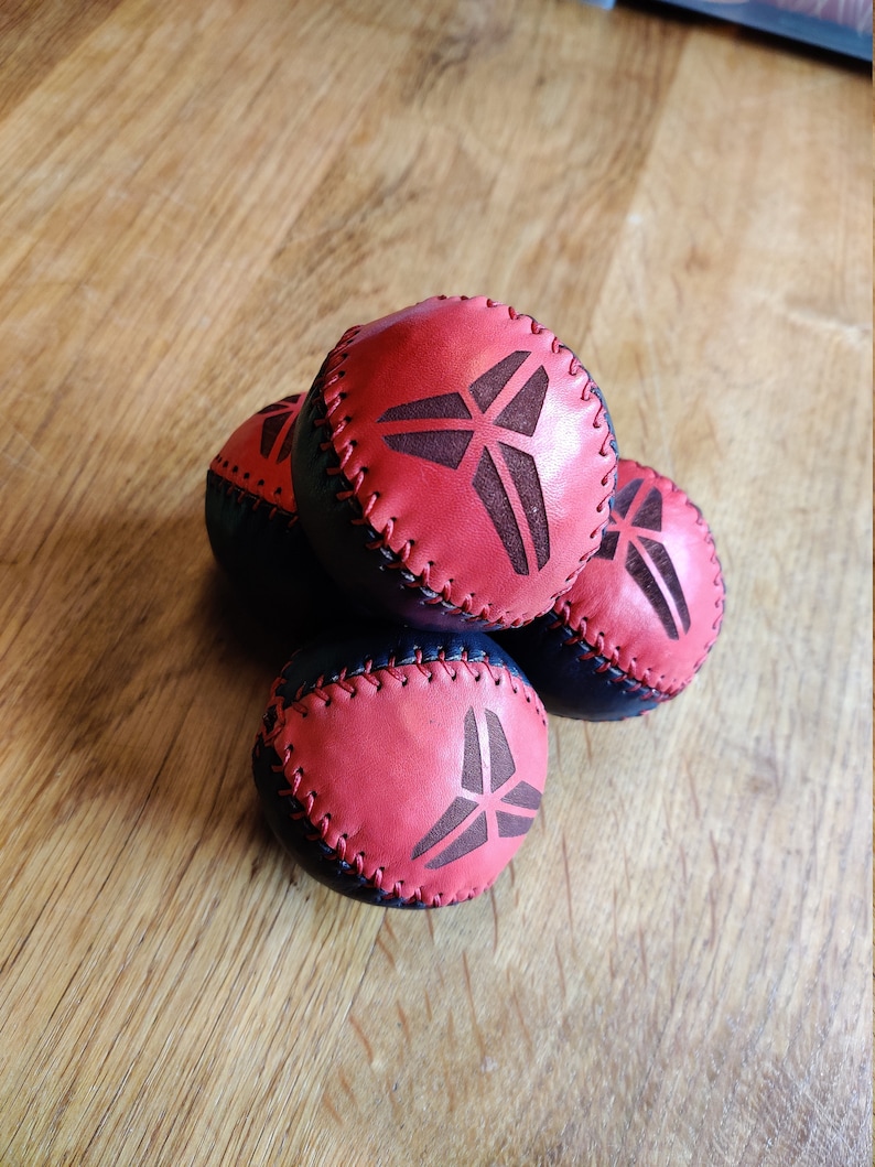 May include: Four leather juggling balls with a black and red design. Each ball has a stitched seam and a black logo of a stylized triangle with a line through it.