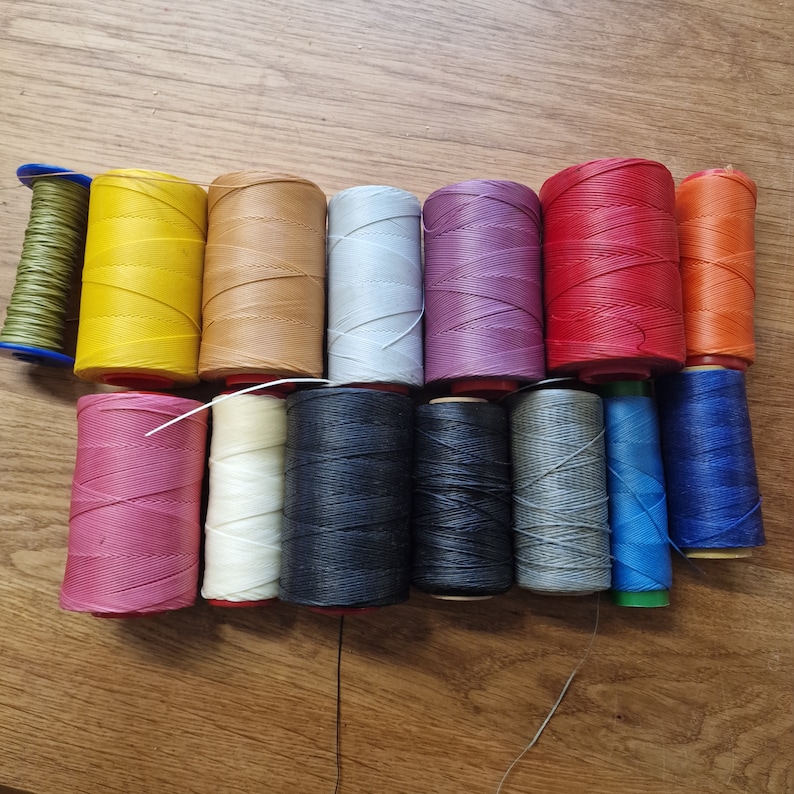 May include: Assortment of 16 spools of waxed thread in various colors, including red, blue, green, yellow, white, black, and pink. The spools are arranged in two rows on a wooden surface.
