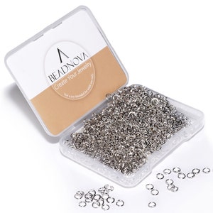 Puede incluir: Un recipiente de plástico transparente lleno de anillos de salto plateados. El recipiente tiene una etiqueta que dice "Beadnova Create Your Jewelry Not a toy - Intended for adult use only".