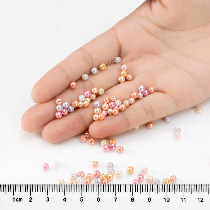 May include: A collection of small, round, faux pearl beads in shades of pink, peach, white, and lavender. The beads are scattered and clustered, with some resting on a surface next to a ruler showing measurements in centimeters up to 12 cm.
