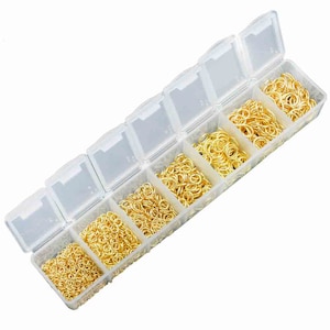 Puede incluir: Una caja de almacenamiento de plástico transparente con seis compartimentos llenos de anillos de salto dorados de varios tamaños.