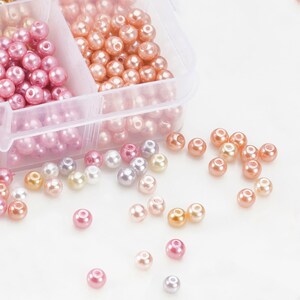 May include: A clear plastic organizer filled with various shades of pink and peach-colored beads. Scattered around the organizer are additional beads in shades of pink, peach, pearl, and silver. These beads are likely used for jewelry making or crafting.