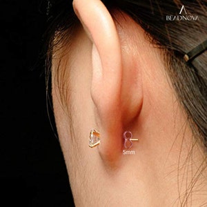 May include: Close-up of a person's ear with a small, clear gemstone earring. The earring is 5mm in diameter and has a gold setting.
