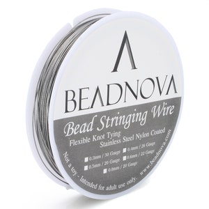 Puede incluir: Un carrete de hilo plateado para enhebrar cuentas con una etiqueta blanca que dice "BEADNOVA Bead Stringing Wire Flexible Knot Tying Stainless Steel Nylon Coated Not a toy - Intended for adult use only. www.beadnova.com 0.3mm / 30 Gauge 0.4mm / 26 Gauge 0.5mm / 20 Gauge 0.6mm / 22 Gauge 0.8mm / 20 Gauge"