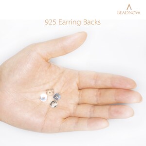 May include: A close-up of a hand holding four silver earring backs. The earring backs are small and round, with a small hole in the center. The text "925 Earring Backs" is visible in the image.
