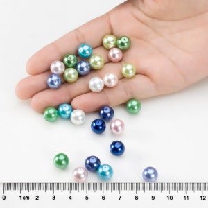 May include: A collection of round, smooth, multicolored beads in shades of blue, green, pink, and white. The beads are shown alongside a ruler, indicating they measure approximately 1 cm in diameter. Each bead features a small hole for threading.