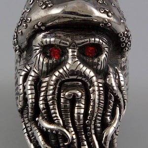 May include: A silver ring with a detailed design of a pirate's face with red eyes and a long beard. The beard is made up of tentacles, giving the pirate a monstrous appearance.