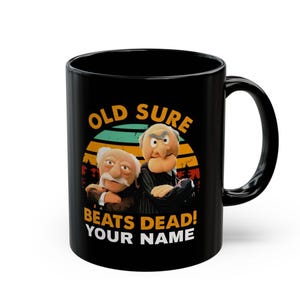 May include: Black ceramic mug featuring a colorful graphic of two Muppet characters with the text "OLD SURE BEATS DEAD! YOUR NAME". The design includes a retro sunset and forest silhouette.