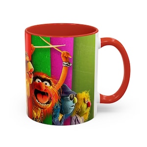 May include: White ceramic mug with a red interior and handle. The mug features a colorful image of the Muppets' Animal and other characters. The image includes Animal holding drumsticks, with a green, pink, and yellow background.