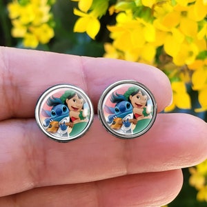 Puede incluir: Un par de pendientes redondos plateados. Cada pendiente presenta una imagen de dibujos animados colorida de Lilo y Stitch. Stitch toca el ukelele. El fondo es de un tono rosado. Los pendientes se sostienen contra un fondo borroso de flores amarillas.