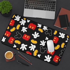 May include: Black rectangular mousepad featuring a pattern of Mickey Mouse elements. The design includes white gloves, red shorts, yellow shoes, and Mickey's head outline. A white mouse sits on the pad, with a laptop and notebook in the background.