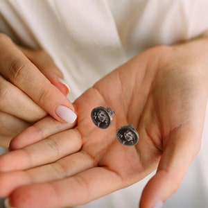 Puede incluir: Un par de pendientes de bot&oacute;n plateados. Cada pendiente presenta una imagen en blanco y negro de una persona. Los pendientes son redondos y tienen cierre de poste. Los pendientes se muestran en una mano abierta.