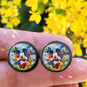 May include: Two round, silver-toned stud earrings. Each earring features a colorful image of Mickey Mouse, Donald Duck, Goofy, and Pluto against a blue sky background. The earrings are held in a hand, with yellow flowers in the background.