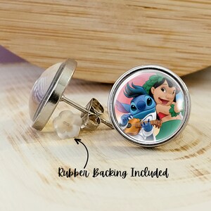 Puede incluir: Un par de pendientes de bot&oacute;n redondos con un engaste de metal plateado. Un pendiente presenta una imagen de dibujos animados de Stitch y Lilo. El otro pendiente tiene un dise&ntilde;o transparente y abovedado. El texto "Rubber Backing Included" es visible.