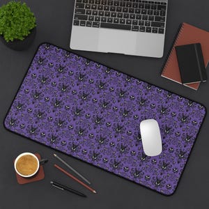 May include: A purple desk mat with a black border featuring a repeating pattern of stylized haunted house and eye designs. A white computer mouse sits on the mat, next to a laptop and other office supplies.