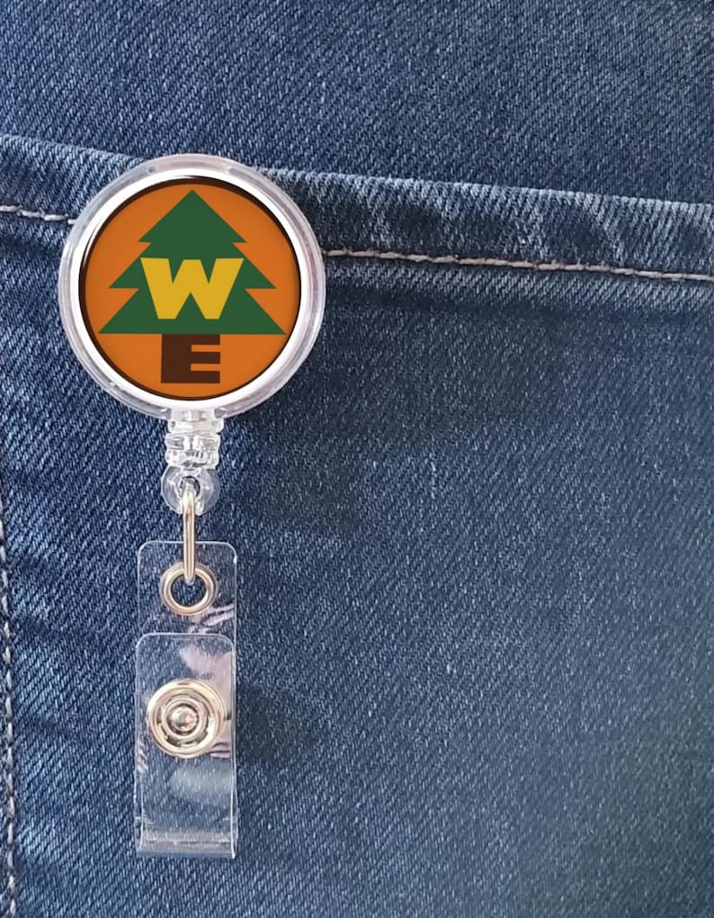 May include: A clear plastic badge holder with a round, colorful button featuring a green pine tree with a yellow "W" and "E" on a brown background.