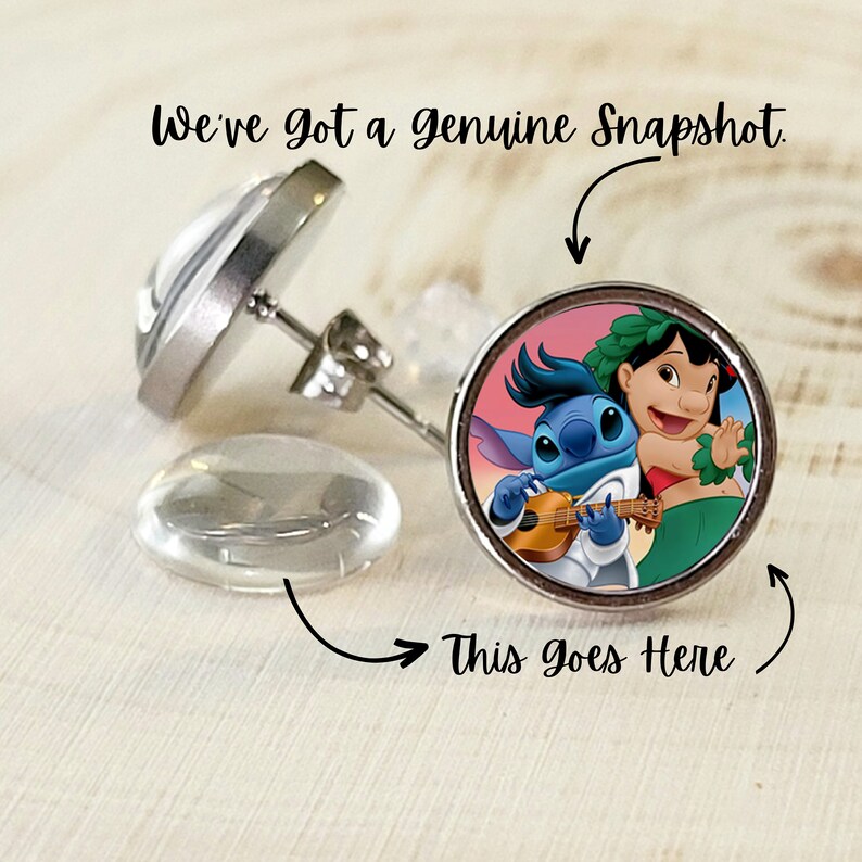 Puede incluir: Pendientes de bot&oacute;n con una imagen de dibujos animados de Lilo y Stitch. Los pendientes tienen un engaste de metal plateado y un reverso transparente y redondeado. El texto "We've got a genuine Snapshot" est&aacute; arriba.