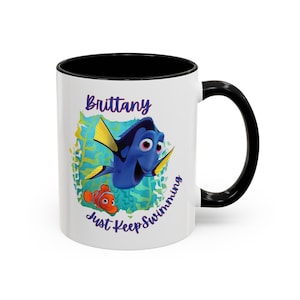 May include: White ceramic mug with a black rim and handle. The mug features a blue and yellow Dory character from the movie Finding Nemo, along with a small orange and white Marlin character. The text "Brittany Just Keep Swimming" is printed on the mug.