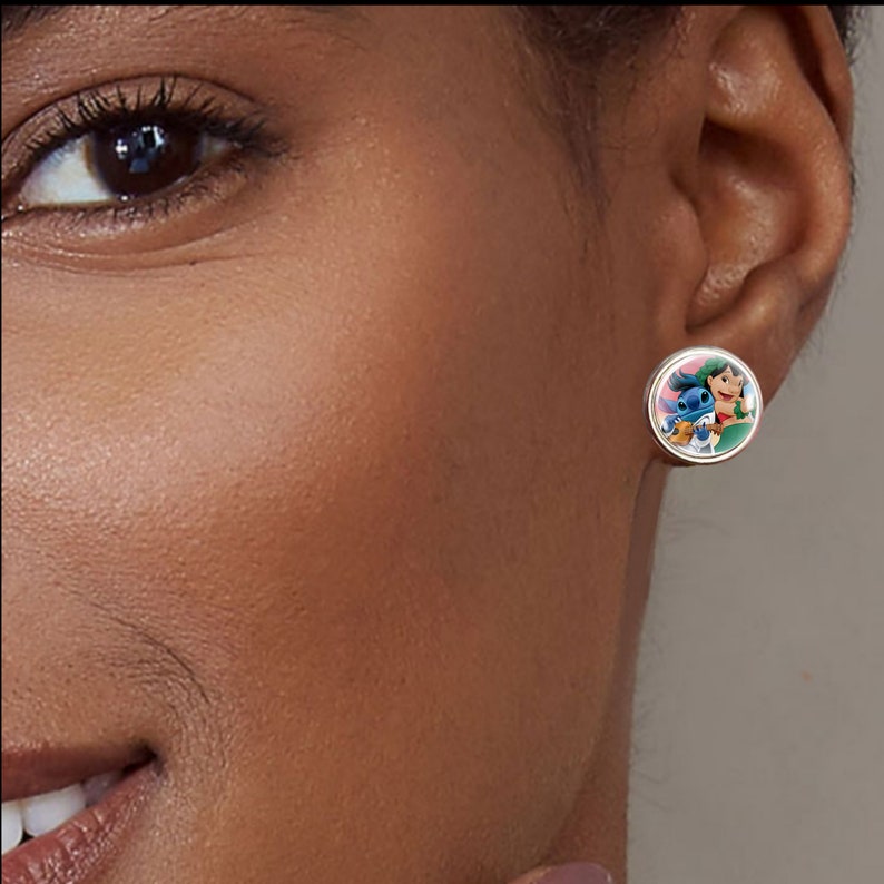 Puede incluir: Pendiente de bot&oacute;n redondo con una imagen de dibujos animados de un extraterrestre azul y una ni&ntilde;a. El pendiente tiene un borde de metal plateado y un fondo rosa. El pendiente se lleva en la oreja de una persona.