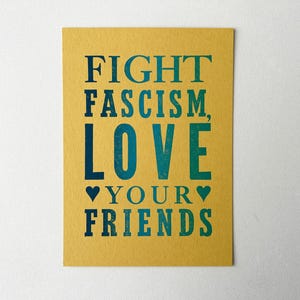Puede incluir: Una tarjeta amarilla con el texto "FIGHT FASCISM, LOVE YOUR FRIENDS" en letras azules y verdes. Dos pequeños corazones están a cada lado de la palabra "YOUR".