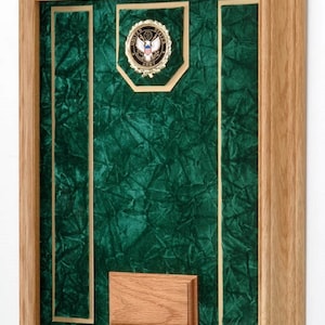 May include: A wooden frame with a green velvet background and a gold-colored United States Army seal. There is a wooden plaque at the bottom of the frame.