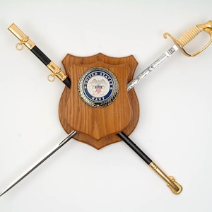 May include: Two crossed swords mounted on a wooden plaque with a United States Navy seal. The swords have gold handles and black and silver blades.