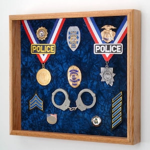 May include: A wooden shadow box display case with a blue velvet background. The case contains a variety of police badges, medals, and other insignia, including handcuffs, a US flag patch, and a pair of blue and gold chevron patches.