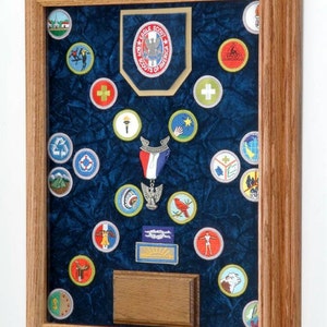 May include: A framed display of Boy Scout patches and insignia. The patches are arranged on a blue velvet background within a wooden frame. The display includes an Eagle Scout emblem and various merit badges.