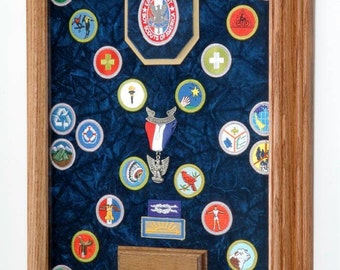 Può includere: Un'esposizione incorniciata di toppe e insegne scout. Le toppe sono disposte su uno sfondo di velluto blu all'interno di una cornice di legno. L'esposizione comprende un emblema Eagle Scout e vari distintivi al merito.