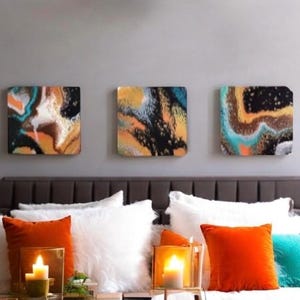 May include: Three abstract paintings with a black, white, brown, orange, and blue color scheme. The paintings are hung on a gray wall above a brown headboard with white and orange pillows.
