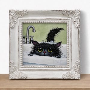 May include: A framed painting of a fluffy black cat with large yellow eyes peeking over the edge of a white bathtub filled with bubbles. A silver faucet is visible in the background against a light green wall. The frame is white and ornate.