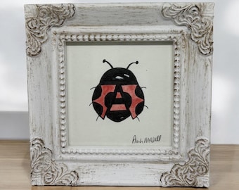 May include: A square, distressed white wooden frame with ornate corner detailing holds a drawing of a ladybug with a black body and red wing accents. The letter A is integrated into the center of the ladybug. The artist signature Ann Marell is visible in the bottom right corner.