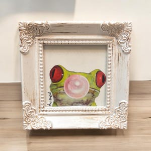 May include: A square, distressed white ornate frame containing a painting of a green frog with large red eyes blowing a pink bubblegum bubble. The artwork features a signature in the bottom left corner and is set against a plain background.