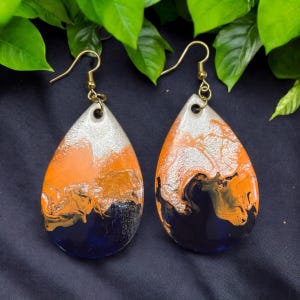 May include: A pair of teardrop-shaped earrings with a marbled design in shades of orange, black, silver, and blue. The earrings have gold hooks.