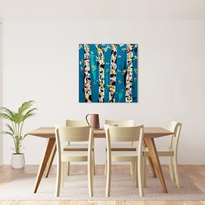 May include: Abstract painting of birch trees with white bark and colorful accents against a teal blue background. The painting is framed and hanging on a white wall above a wooden dining table with four chairs.