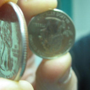 May include: Two United States quarter coins are held between fingers. One coin is viewed from the side, showing the reeded edge. The other coin is viewed from the front, displaying the image of George Washington.